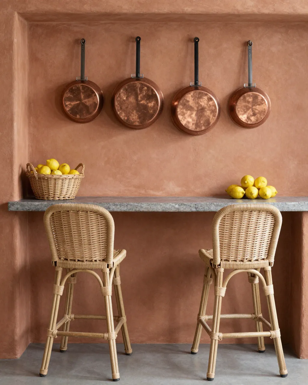 A small kitchen nook has terracotta walls, hanging copper pans, rattan stools and bowls of lemons.