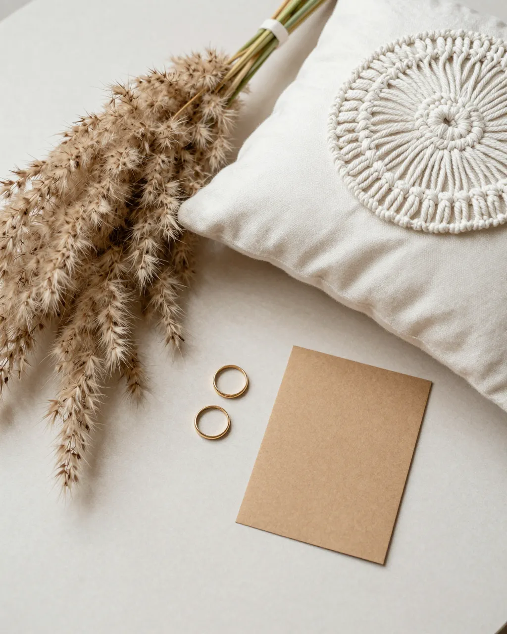 Flat lay arrangement of dried pampas grass, macrame ring pillow, gold wedding bands, and blank kraft card on a soft neutral