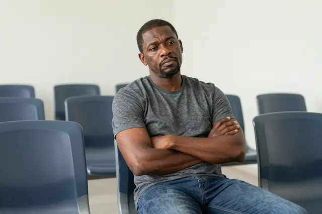 A middle aged Black man sits with folded arms among empty plastic waiting room chairs looking bored