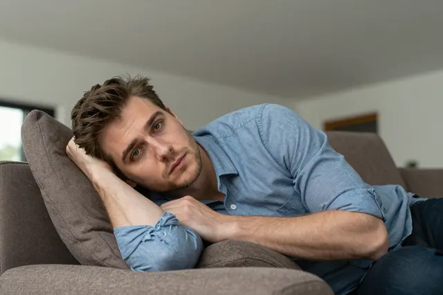 A bored man in his thirties reclines on a brown sofa resting his head on his hand