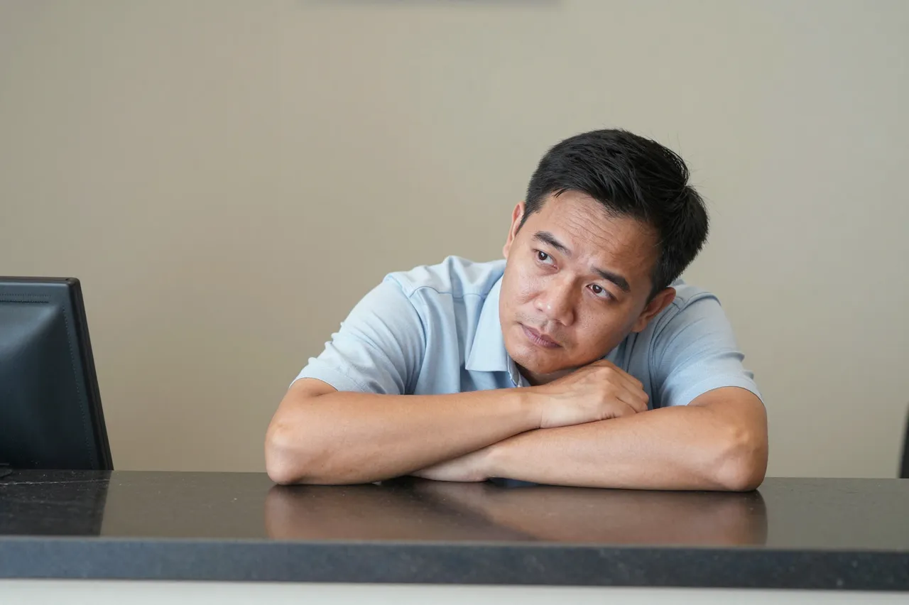 Bored middle aged Southeast Asian man rests his chin on folded arms at a dark counter desk