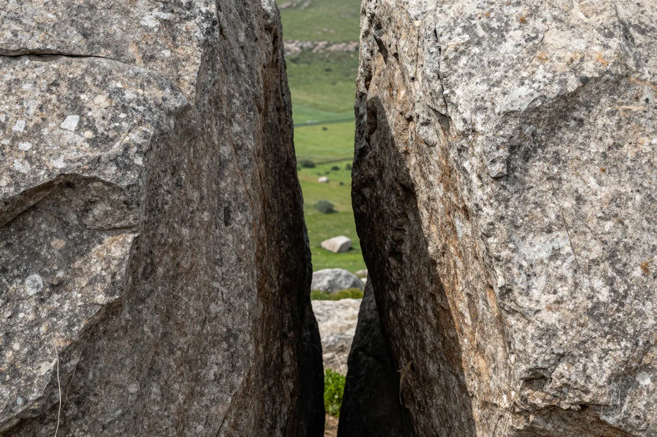 A narrow gap between two large granite boulders frames a view of a sunlit rolling green valley.