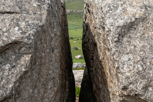 A narrow gap between two large granite boulders frames a view of a sunlit rolling green valley.