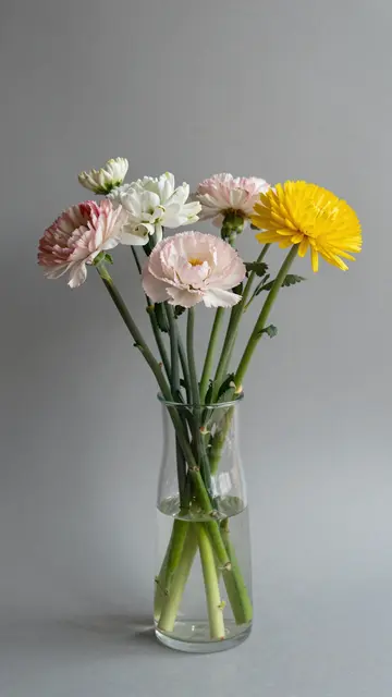Mixed pink, white and yellow flowers arranged in a clear glass vase against a plain gray background