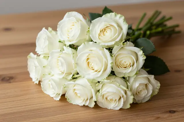 A fresh bouquet of white roses rests flat on a light brown wooden table.