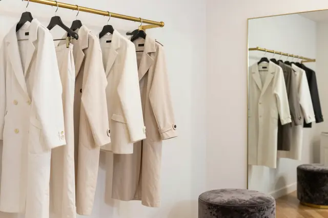 Neutral toned outerwear hangs on a brass clothing rack next to a tall gold framed mirror and dark velvet ottoman.
