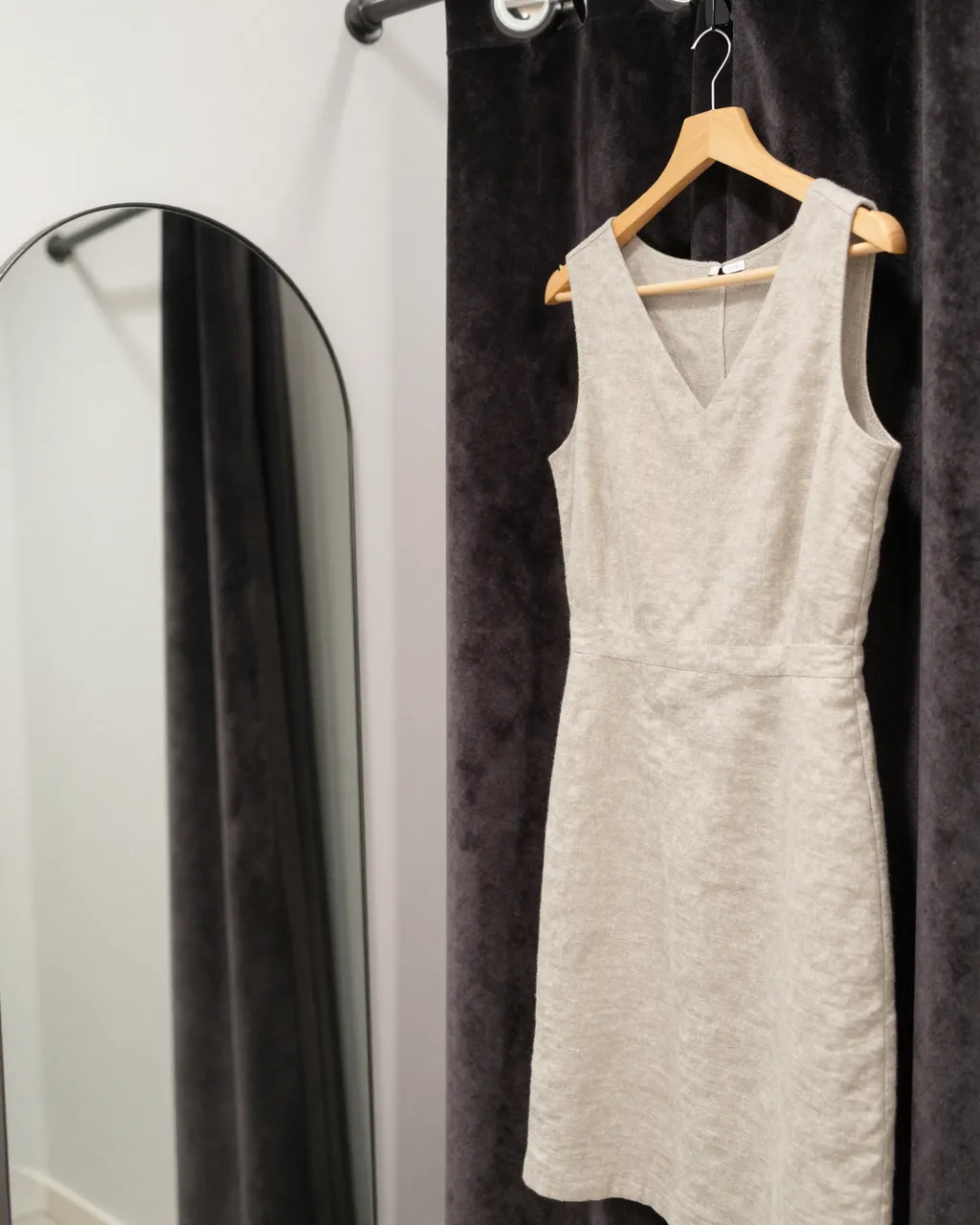 A plain linen dress hangs on a wooden hanger next to a velvet fitting room curtain and arched mirror.