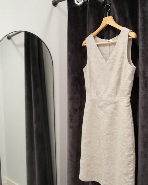 A plain linen dress hangs on a wooden hanger next to a velvet fitting room curtain and arched mirror.