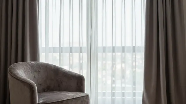 Plush gray velvet armchair sits beside a tall window with sheer and dark drapes