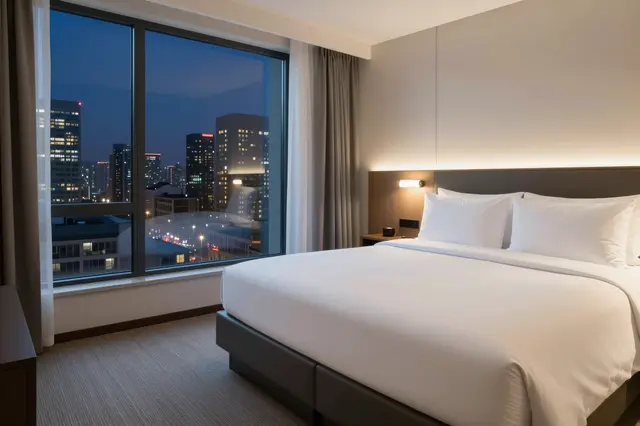 Modern hotel room with a king bed overlooking lit city skyline at dusk through a large window
