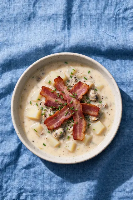 Overhead view of speckled bowl of clam chowder topped with bacon, chives and pepper on blue linen