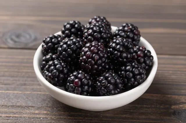 A small white bowl filled with ripe blackberries sits on a dark wooden table.