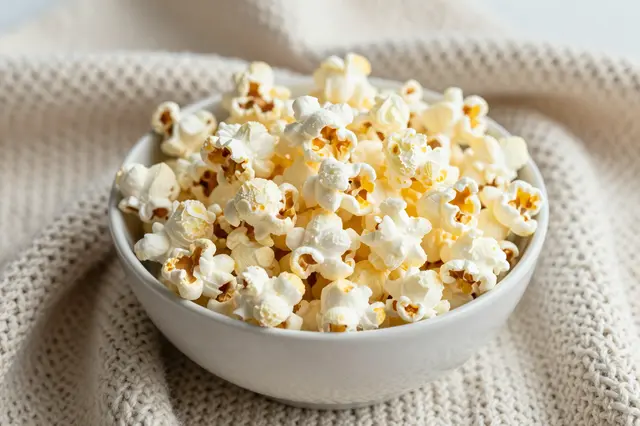 A white bowl of buttery popcorn rests on a soft beige knitted blanket.