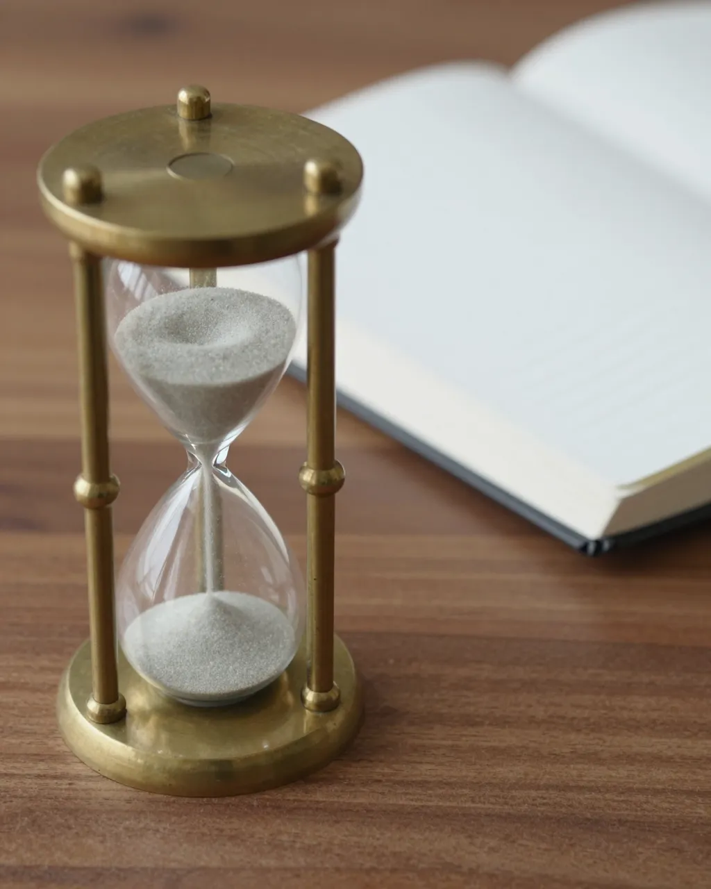 Brass hourglass with running pale sand sits next to an open notebook on a walnut desk.