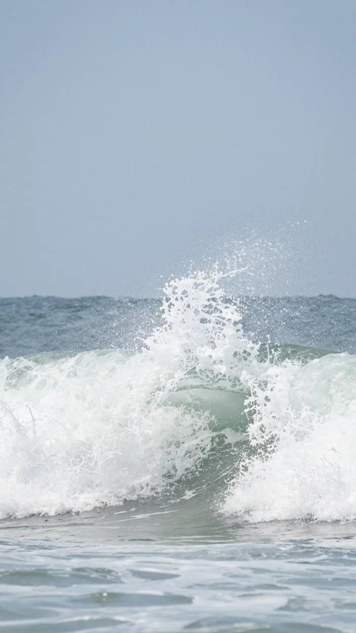 A breaking ocean wave erupts into white foam under a pale clear blue sky