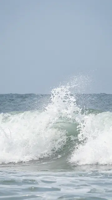 A breaking ocean wave erupts into white foam under a pale clear blue sky