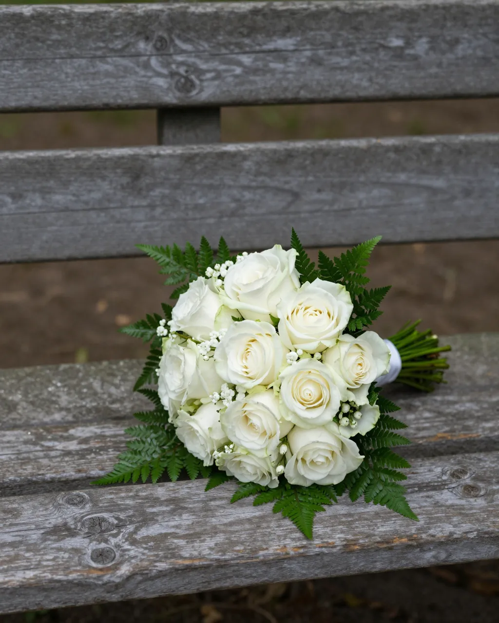 A bridal bouquet of white roses and ferns rests on a weathered wooden park bench in soft shade.