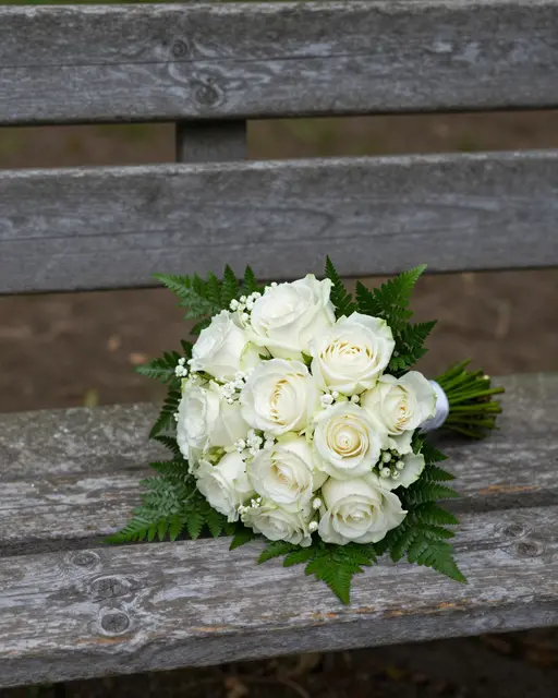 A bridal bouquet of white roses and ferns rests on a weathered wooden park bench in soft shade.