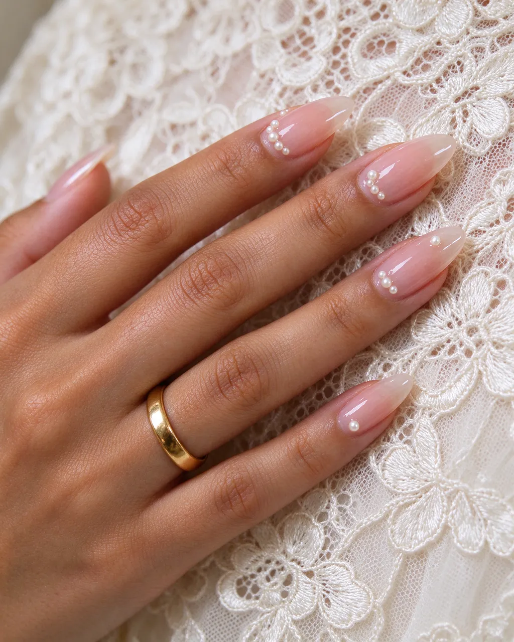 Close-up of a hand with sheer pink pearl almond nails and a plain gold band resting on ivory lace fabric