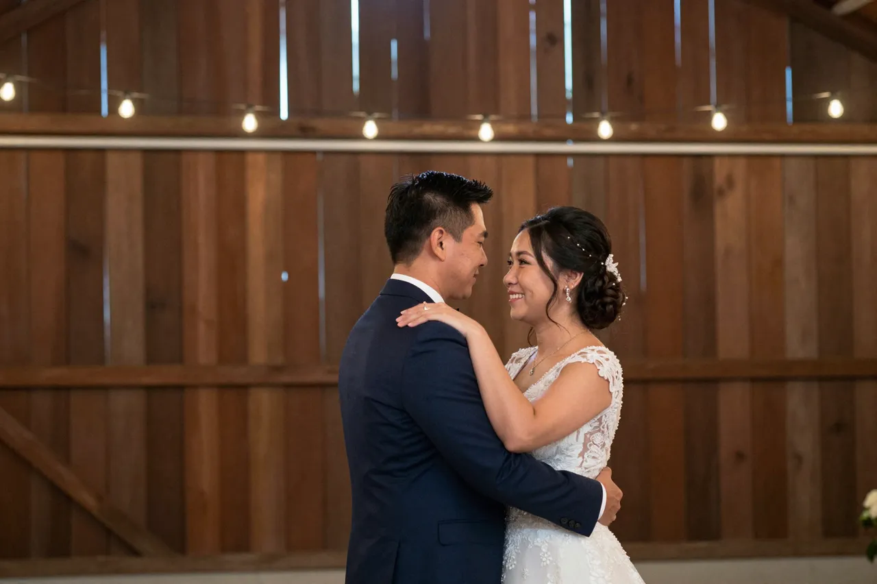 A Southeast Asian bride and groom dance closely facing each other inside a wooden barn under string lights