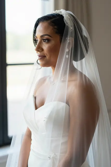 Mixed-race bride in her thirties stands by a window, soft morning light catching her beaded veil.