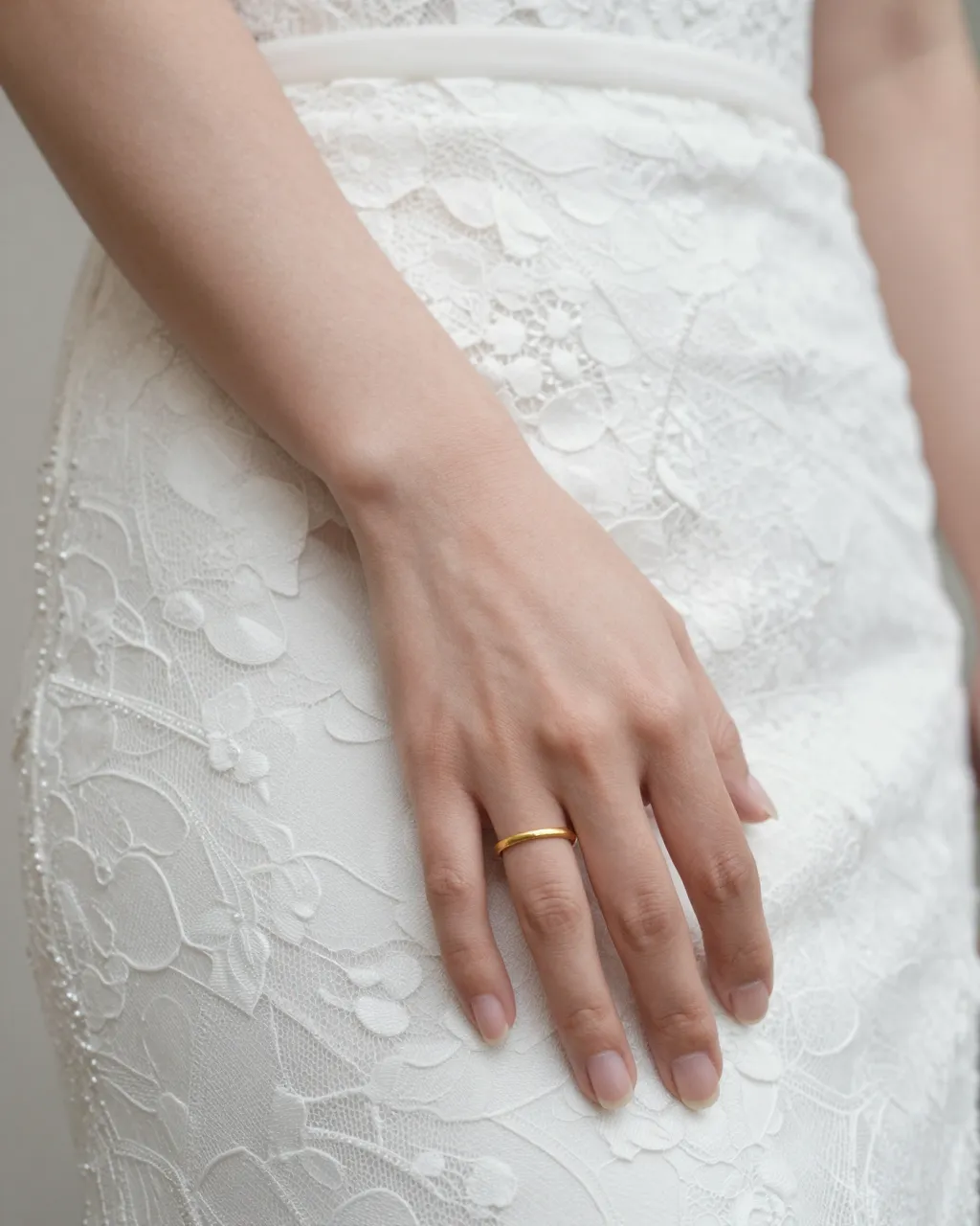 Close-up of a bride's hand with a thin gold wedding band resting on white lace bridal dress