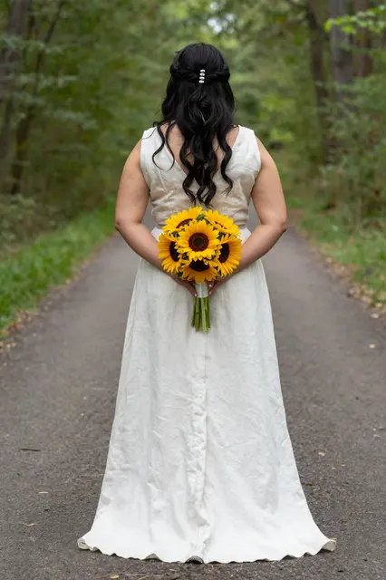 Back view of a bride in a linen gown holding a sunflower bouquet behind her on a wooded path