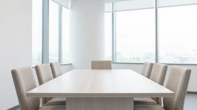 Light wood conference table with beige linen chairs sits in a sunlit boardroom with tall windows