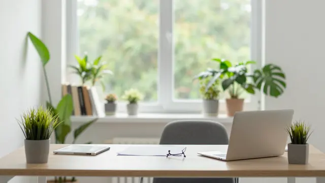 A light wooden home office desk with a laptop, plants, and natural light from a large window.