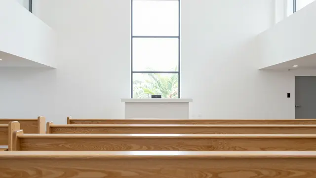 Rows of pale wooden pews face a plain altar below a tall window in a bright empty modern church.