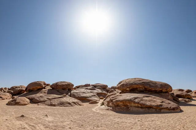 Bright sun with starburst effect shines over weathered boulders on sandy desert under a clear cloudless blue sky