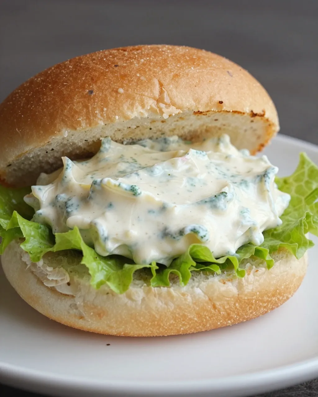 A close-up of a brioche bun sandwich with lettuce and herb-speckled creamy remoulade on a white plate.