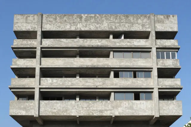 Upper floors of a weathered brutalist concrete building stand against a clear bright blue sky.
