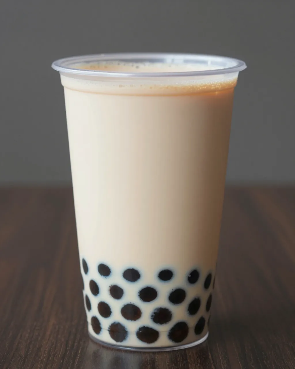 A clear plain plastic cup of milk bubble tea with tapioca pearls sits on a dark wooden table