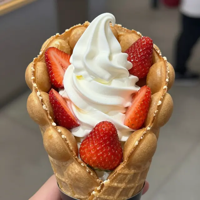 A hand holds a bubble waffle cone topped with vanilla soft serve and fresh strawberries.