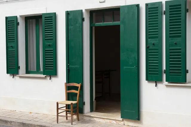 Wooden chair sits beside an open doorway on a white building with green wooden shutters at midday.