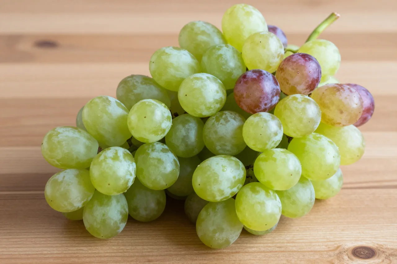A cluster of mostly green grapes with scattered red berries rests on a light wooden tabletop.