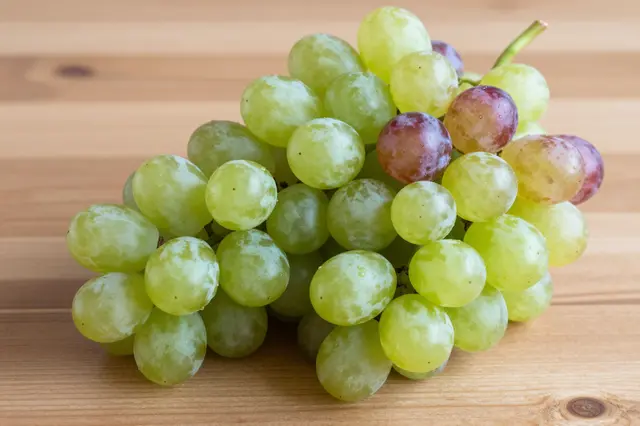 A cluster of mostly green grapes with scattered red berries rests on a light wooden tabletop.