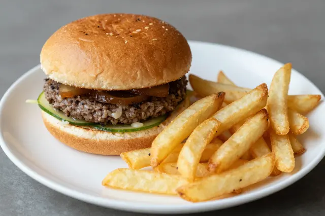 A freshly prepared burger and pile of thick cut french fries rest on a white plate atop a gray tabletop.