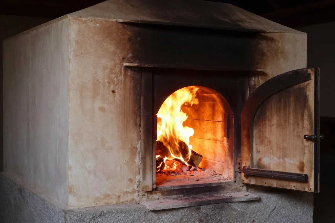 An open wood-fired kiln has bright flames licking out of its firebox door in low ambient light