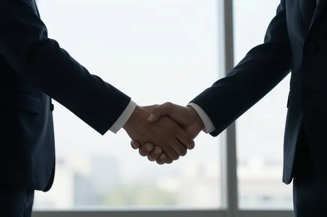 Two people in dark business suits shake hands in front of a bright blurred office window