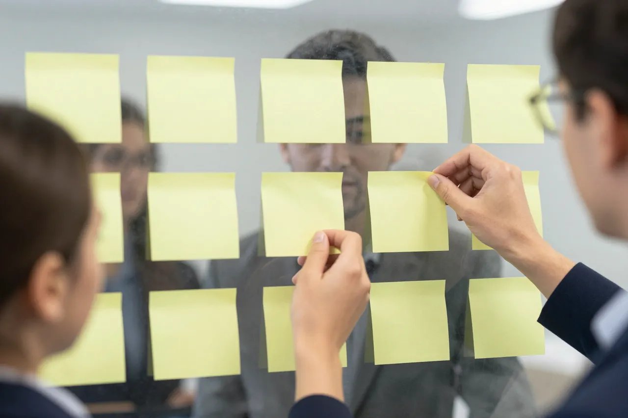 Office workers place blank yellow sticky notes on a glass partition during a collaborative planning session.