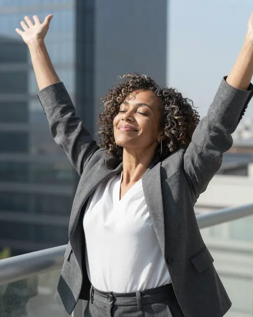 A mixed-race woman in a business suit raises her arms with eyes closed on a sunny office balcony