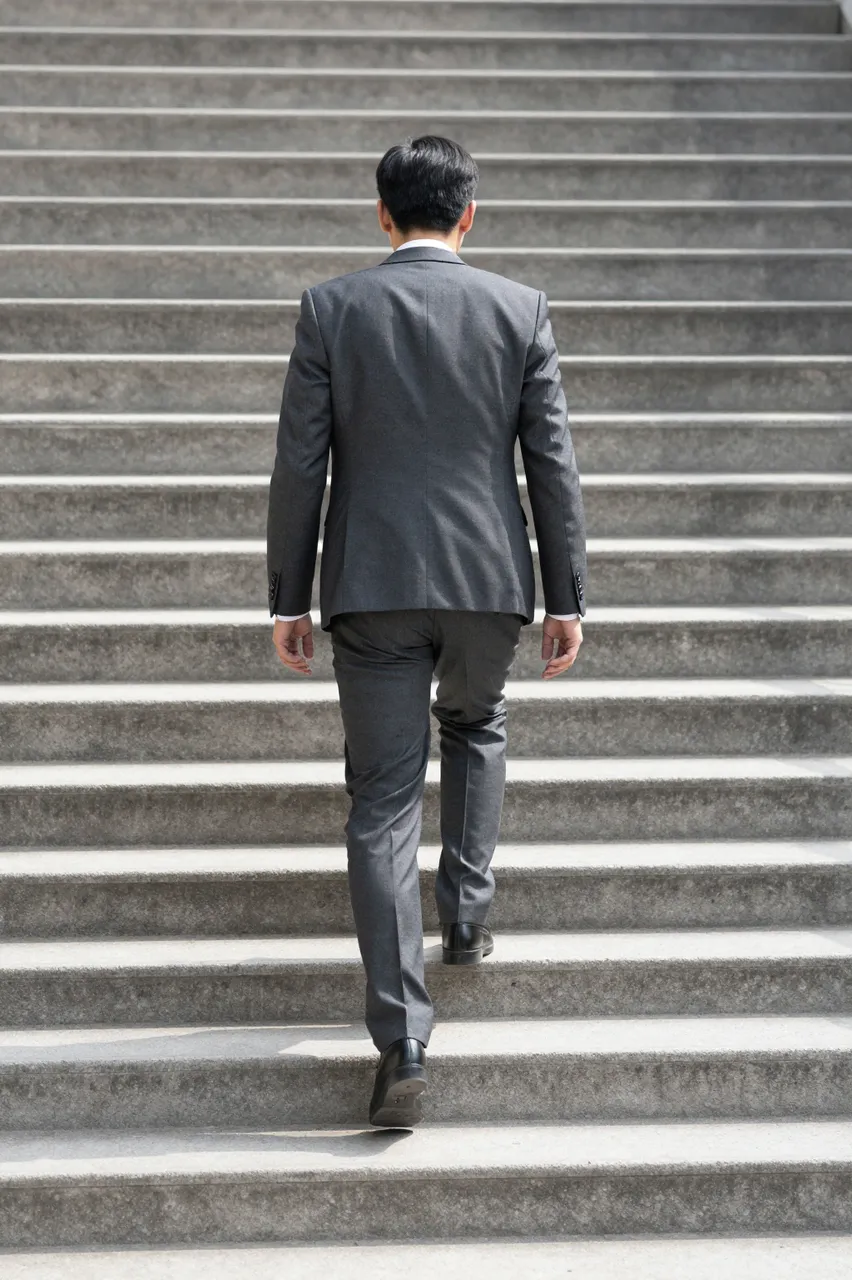 A businessman seen from behind walks up a sunlit outdoor concrete staircase.