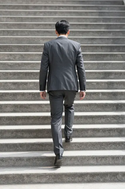 A businessman seen from behind walks up a sunlit outdoor concrete staircase.