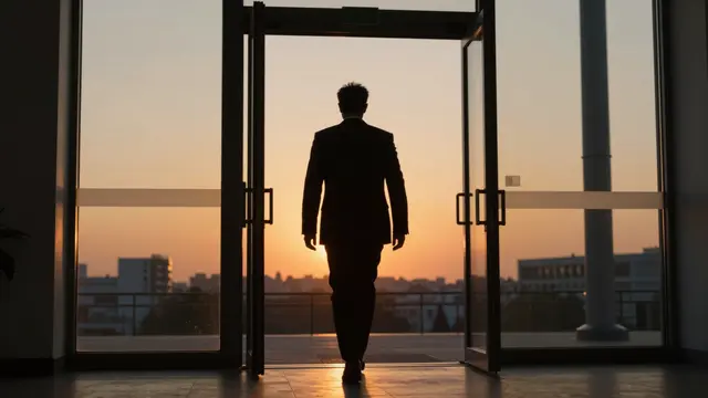 Silhouette of a suited businessman walking through open office exit doors toward an orange sunset