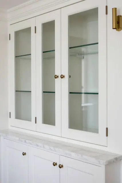 Empty glass-front white pantry cabinets with marble countertop and brass sconce on a plain wall