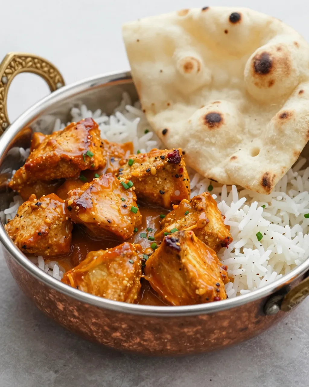 Copper serving bowl holds butter chicken curry over basmati rice with a piece of charred naan bread.