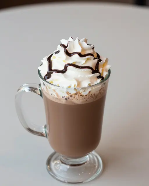 Clear glass mug of cafe mocha topped with swirled whipped cream and chocolate syrup drizzle on a light table