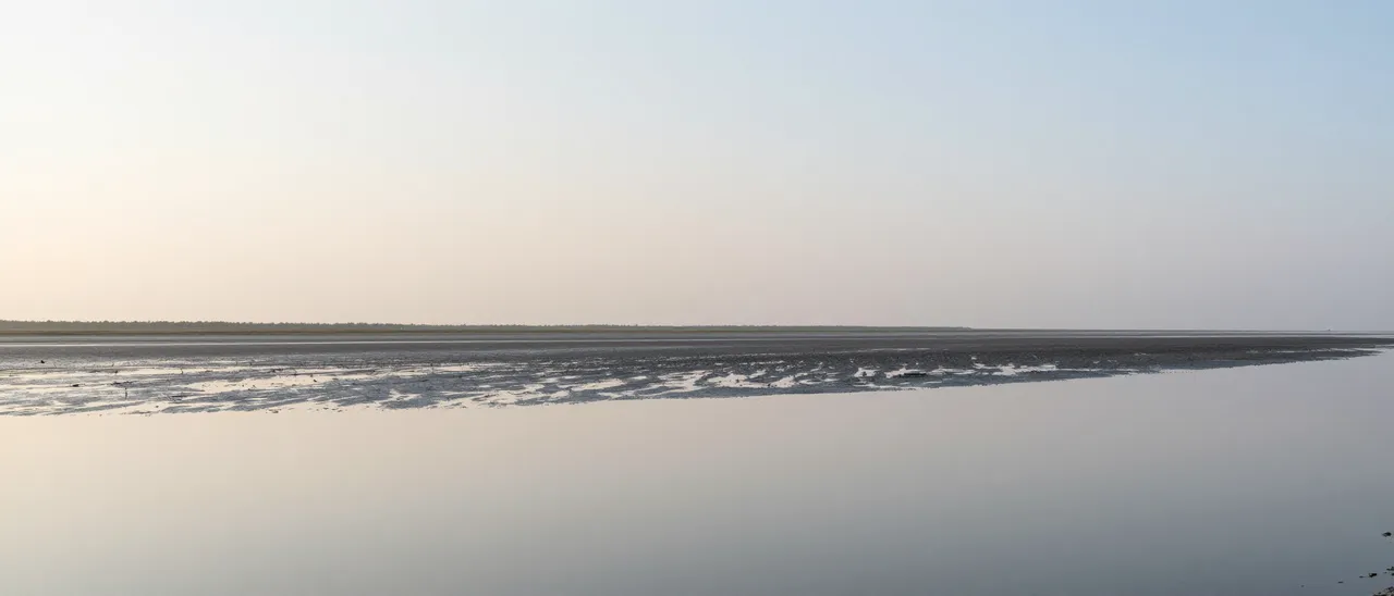 A calm tidal flat reflects the pale soft gradient of the early morning sky beside a distant narrow land strip